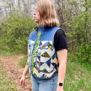 May include: A sleeveless waistcoat featuring a blue denim upper section and a lower section constructed from a multicoloured patchwork quilt pattern of triangles. The garment includes a high collar, a front button closure, and two large patch pockets.
