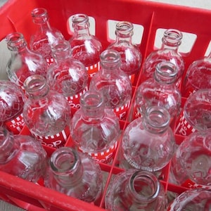 May include: A red plastic crate filled with clear glass bottles. The bottles have red and white labels with the text "The Pop" printed on them.