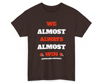 May include: A dark brown short-sleeve t-shirt featuring the orange and white text WE ALMOST ALWAYS ALMOST WIN, accompanied by two small orange football helmet graphics and the white text CLEVELAND FOOTBALL centred on the front chest area.