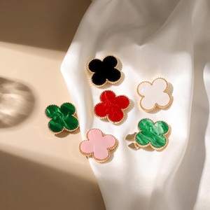 May include: A collection of six four-leaf clover charms with gold-toned beaded edges. The charms feature smooth, solid, or marbled surfaces in black, white, red, light pink, and two shades of green. Each clover has a distinct, rounded petal shape.