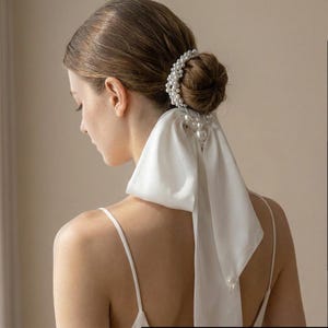 May include: A white satin hair accessory with a pearl accent. The accessory is tied in a bow and is worn in the hair. The pearls are white and round. The hair accessory is a bridal or formal wear item.