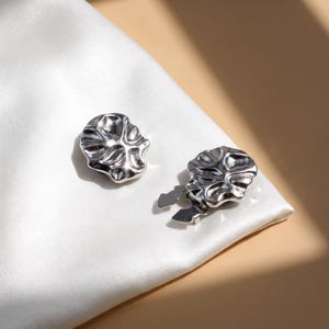May include: A pair of silver-toned metal earrings featuring a rounded, organic, and irregular shape with a high-shine, reflective surface. The design includes a textured, rippled finish and a secure clip-on mechanism on the back of each piece.