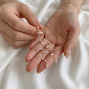 Puede incluir: Pendientes colgantes de oro rosa con un delicado diseño de hojas. Cada pendiente tiene una barra delgada con pequeñas piedras claras en forma de lágrima. Los pendientes se muestran sobre un fondo de tela blanca.