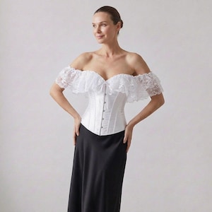 May include: A white corset with a sweetheart neckline and delicate lace trim. The off-the-shoulder design features ruffled lace sleeves. The corset is paired with a long, black skirt, creating an elegant ensemble.