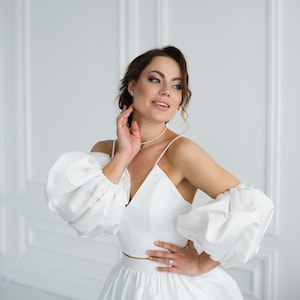 May include: A white two-piece bridal outfit with a fitted top and a full skirt. The top has a V-neckline and large, puffy sleeves. The model is wearing a pearl necklace.