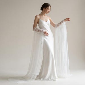 Peut inclure: Une robe de mariée en satin blanc avec des bretelles spaghetti et une traîne fluide. La robe présente de longues manches transparentes qui se prolongent en un design de type cape. Les manches sont ornées de volants horizontaux délicats.