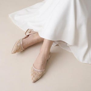 Könnte beinhalten: Beigefarbene Ballerinas mit spitzer Zehenpartie, verziert mit zarten Perlen und Perlen. Die Schuhe haben einen transparenten Mesh-Einsatz und einen Knöchelriemen. Die Schuhe werden mit einem weißen Kleid getragen.