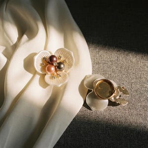 Puede incluir: Dos broches en forma de flor con pétalos blancos y un centro de tres perlas en marrón oscuro, marrón claro y gris, acentuados con pequeños cristales transparentes. Un broche muestra el frente y el otro la parte posterior de metal dorado.