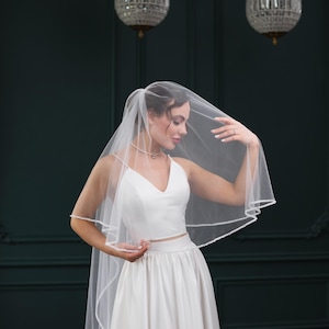 Könnte beinhalten: Eine Braut in einem weißen Brautkleid mit einem langen, weißen Schleier. Der Schleier ist mit einer zarten Spitze verziert. Die Braut schaut auf den Schleier.