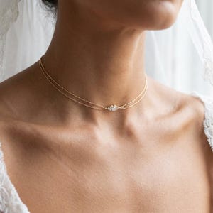 Puede incluir: Un delicado collar de oro con dos finas cadenas. El collar presenta un diseño central y simétrico de pequeñas piedras brillantes. La joya se lleva en el cuello de una persona, lo que sugiere que es una pieza de novia o de vestir.