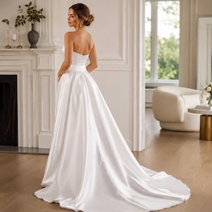 May include: A strapless white satin wedding dress featuring a fitted bodice with a defined waistline and a full, floor-length skirt that extends into a train. The smooth, lustrous fabric drapes elegantly from the waist, creating a voluminous silhouette.