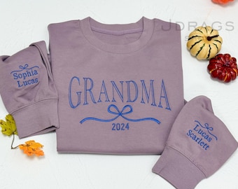 May include: A mauve sweatshirt with the word "GRANDMA" embroidered in blue, along with a bow and the year "2024". Sleeves feature embroidered names. Decorative pumpkins and leaves are in the background. A cozy, personalized gift.