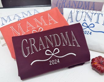 May include: Folded sweatshirts in various colors, including coral, burgundy, and white, are displayed. Each sweatshirt features embroidered text: "MAMA," "GRANDMA," and "AUNTIE." The year "2024" is embroidered on some. A decorative pumpkin is in the foreground.