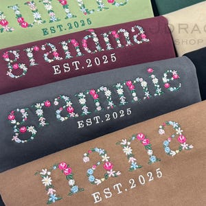Peut inclure: Gros plan de plusieurs sweat-shirts de différentes couleurs, chacun brodé d'un motif floral avec le nom et "EST. 2025". Les noms incluent "Grandma", "Grammie" et "Nana", avec des lettres florales roses, blanches et bleues.