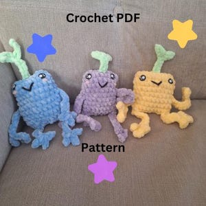 Op de afbeelding: Drie gehaakte pluche speelgoedjes in de vorm van een wezen met een groene stengel erop. De speelgoedjes zijn blauw, paars en geel. De tekst "Crochet PDF" en "Pattern" is zichtbaar in de afbeelding.
