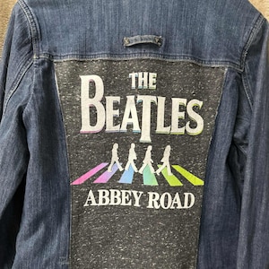 May include: A dark blue denim jacket featuring a graphic on the back with the words "THE BEATLES" in white, and the iconic Abbey Road album cover image. The graphic includes the band members walking on a crosswalk with colorful stripes.