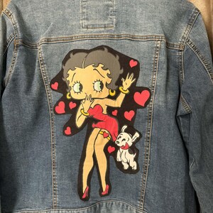 May include: A blue denim jacket featuring a Betty Boop graphic on the back. The graphic shows Betty Boop in a red dress with a small dog, surrounded by red hearts. The jacket has a classic button-up design.