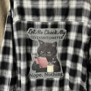 May include: Black and white plaid button-down shirt with a graphic on the back. The graphic features a black cat wearing glasses, reading a book, and holding a mug. Text on the graphic reads "Let Me Check My GIVEASHITOMETER" and "Nope, Nothing."