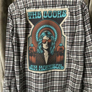 May include: A brown, black, and white plaid flannel shirt with a large patch featuring the band name "THE DOORS" and the name "JIM MORRISON". The patch has a retro design with a portrait of Jim Morrison.