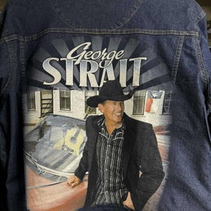 May include: Dark blue denim jacket featuring a graphic of George Strait. The graphic shows George Strait in a black hat and jacket, leaning against a red car. The name "George Strait" is printed in large white letters above the image.