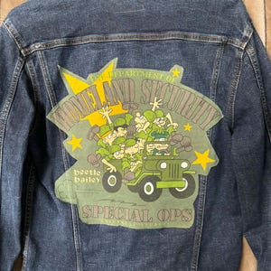 May include: Dark blue denim jacket with a graphic on the back featuring a cartoon of soldiers in a green jeep. The graphic includes the text "THE DEPARTMENT OF HOMELAND SECURITY" and "SPECIAL OPS".