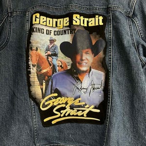 May include: A blue denim jacket with a black and gold patch featuring a photo of George Strait, a country music singer, on a horse. The patch reads "George Strait, King of Country" and includes his signature.