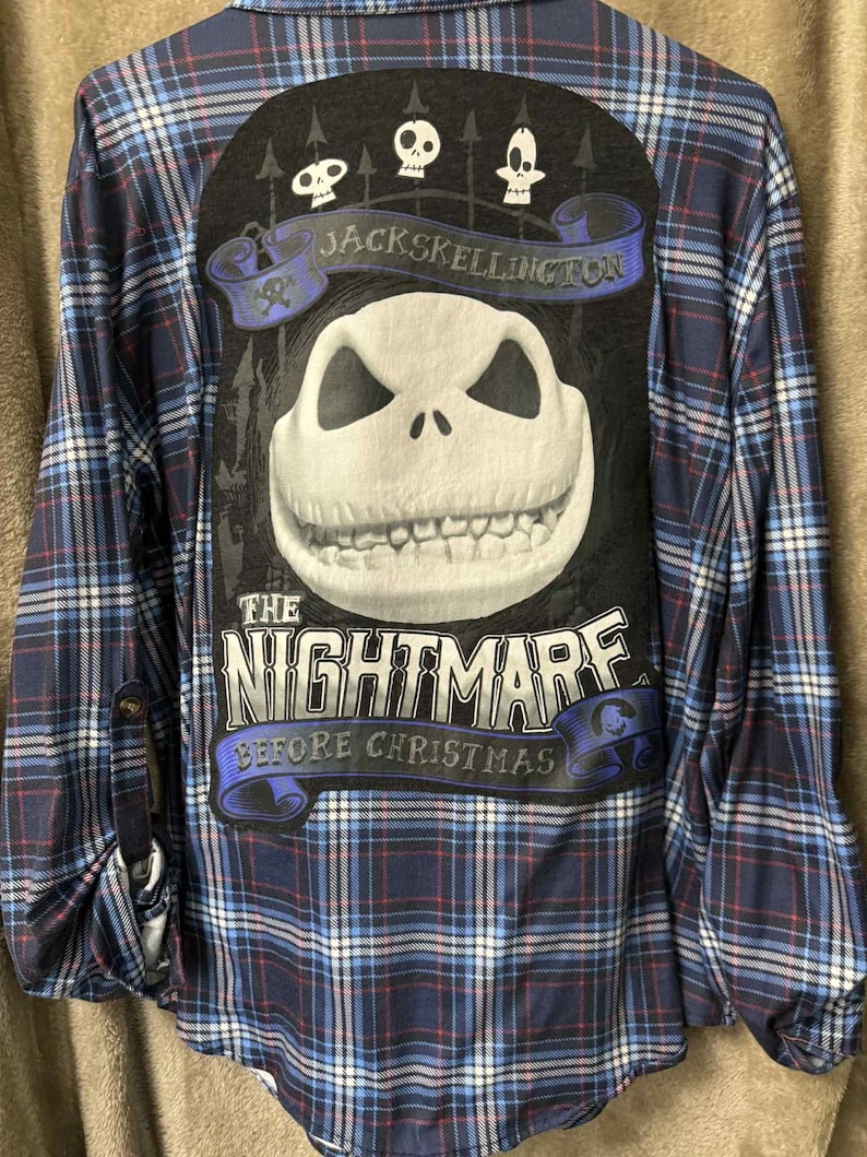 May include: A long-sleeved, button-down flannel shirt in a blue, red, and white plaid pattern. The back features a large graphic of Jack Skellington from "The Nightmare Before Christmas" with the text "Jack Skellington" and "The Nightmare Before Christmas".