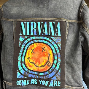 May include: Dark wash denim jacket with a large patch on the back featuring the band name "NIRVANA" in turquoise lettering. The patch includes a smiley face with crossed-out eyes and the phrase "COME AS YOU ARE".
