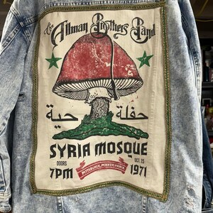 May include: A light blue denim jacket with a patch on the back. The patch features a mushroom illustration, Arabic text, and the words "The Allman Brothers Band", "Syria Mosque", "Oct. 15, 1971", "Doors 7PM", and "Pittsburgh, Pennsylvania".