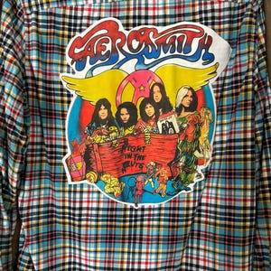 May include: A long-sleeved, button-down shirt with a plaid pattern in blue, red, yellow, and black. The back features a large, colorful graphic of the band Aerosmith, with the band's name and the phrase "Fight in the Ruts."