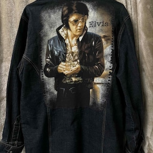 May include: Dark denim jacket featuring a portrait of Elvis Presley on the back. The image shows Elvis in a black leather jacket and sunglasses, with the name "Elvis" printed above. The jacket has a classic button-up design.