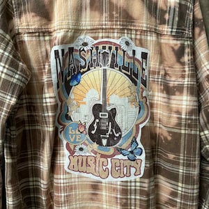 May include: Brown plaid flannel shirt with a distressed design. The back features a colorful patch with the words "Nashville Music City" and a guitar illustration, along with butterflies and flowers.