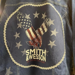 May include: A dark blue denim jacket with a large circular patch on the back. The patch features a silver chain border, a red, white, and blue eagle with outstretched wings, and the text "Smith & Wesson An American Story".