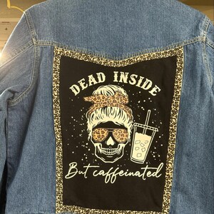 May include: A blue denim jacket with a black patch on the back. The patch features a skull with a leopard print bandana, sunglasses, and a cup of iced coffee. The text on the patch reads "Dead Inside But Caffeinated."