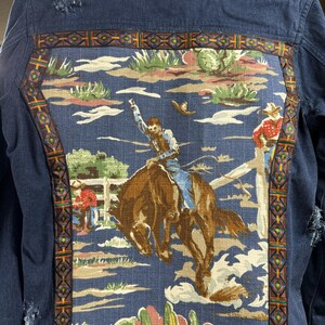 May include: A blue denim jacket with a colorful fabric patch on the back. The patch features a cowboy riding a bucking horse in a western landscape.