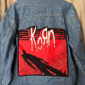 May include: A light blue denim jacket with a large red and black graphic on the back. The graphic features the band name "Korn" in white, with a silhouetted figure. The jacket has a classic button-up design.
