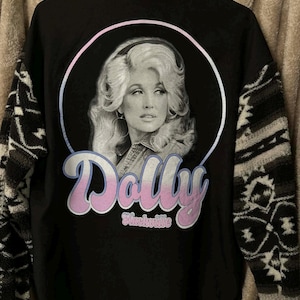 May include: Black long-sleeve shirt featuring a portrait of Dolly Parton inside a circle, with the name "Dolly" in pink script and "Nashville" below. The sleeves have a patterned design in shades of black, white, and gray.