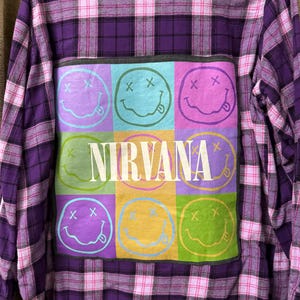 May include: Purple plaid flannel shirt with a Nirvana patch. The patch features a grid of colorful smiley faces with crossed-out eyes and a tongue sticking out, with the band's name in white block letters.