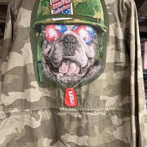 May include: Camouflage jacket featuring a bulldog wearing sunglasses with red, white, and blue lenses. The jacket has a patch with a dog wearing a military helmet and a dog tag that says "USA". The text "Stars & Stripes Winning Fights" is also visible.
