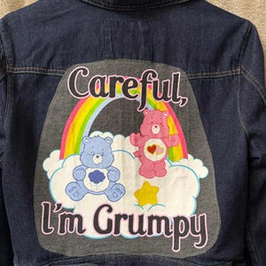 May include: A dark blue denim jacket with a colorful graphic on the back. The graphic features two Care Bears, a rainbow, and the text "Careful, I'm Grumpy". The jacket has a classic collar and button closures.
