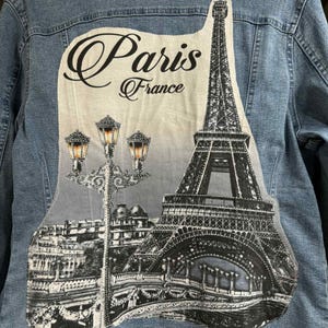 May include: Light blue denim jacket with a large graphic on the back featuring the Eiffel Tower, street lamps, and the words "Paris France" in a decorative font. The graphic is in shades of gray and black.