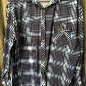 May include: A navy blue plaid button-down shirt with a chest pocket. The shirt features a classic plaid pattern in shades of blue, red, and white. It has a collared neckline and long sleeves. The shirt is buttoned up and appears to be made of a soft fabric.