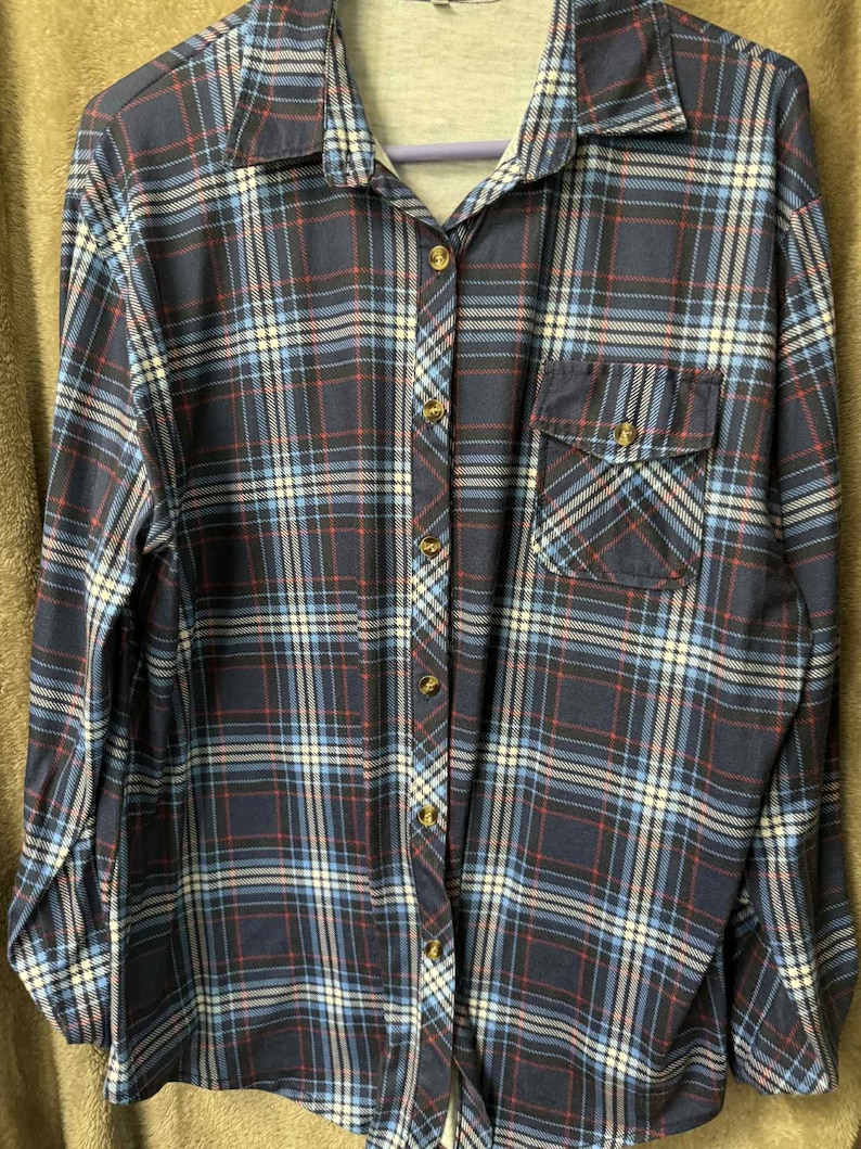 May include: A navy blue plaid button-down shirt with a chest pocket. The shirt features a classic plaid pattern in shades of blue, red, and white. It has a collared neckline and long sleeves. The shirt is buttoned up and appears to be made of a soft fabric.