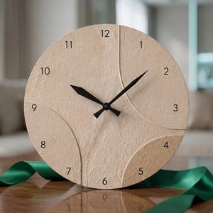 May include: A round wall clock with a textured, light beige face featuring curved embossed lines. The clock has black hour and minute hands and black Arabic numerals from 1 to 12 printed around the perimeter of the circular clock face.