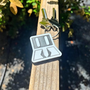 May include: A white and gray geometric design keychain with two keys attached. The keychain is made of a 3D-printed material and is hanging on a wooden post. The keys are silver and black.