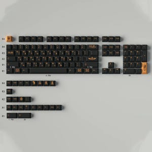 May include: A black mechanical keyboard with gold keycaps featuring various symbols and designs. The keyboard has a number pad and function keys. The keys are labeled with the standard QWERTY layout. The keyboard is on a white surface.