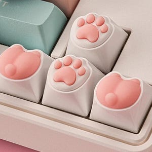 May include: Four white keyboard keycaps with pink paw print and bunny designs. The keycaps are for a mechanical keyboard.