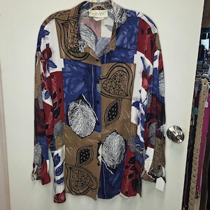 May include: A long-sleeve button-down shirt featuring a multi-colored patchwork pattern of blue, brown, red, and white. The design includes graphic leaf illustrations and abstract shapes. The collar is pointed, and the brand label reads Jessie.