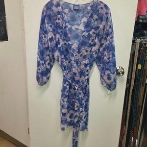 May include: A sheer, lightweight wrap robe featuring a vibrant blue and purple floral pattern. The garment has long, loose sleeves, a deep V-neckline, and a matching fabric tie belt at the waist. The fabric is semi-transparent with a soft drape.