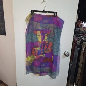 May include: A sheer scarf with a colourful abstract design featuring a figure in a hat. The artwork uses shades of purple, blue, yellow, and red. The word "piceno" is written on the scarf.